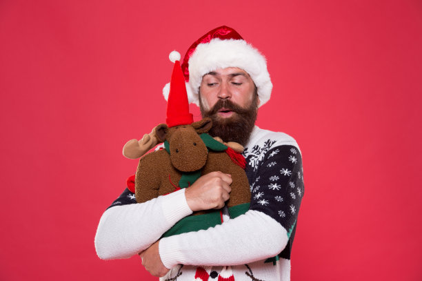 圣诞节准备的概念。有胡须的潮人脸冬季毛衣。寒假。柔软的玩具。拥抱和爱。买礼物。冬季销售。冬天的盛宴。驯鹿。圣诞老人的帽子，手持圣诞礼物红色背景图片下载