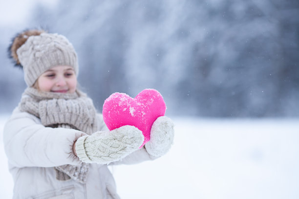 微笑的白人女孩穿着温暖的针织衣服抱着一颗粉红色的心在冬日的雪景背景。图片下载