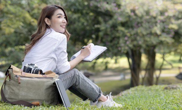 美丽微笑的大学生拿着笔和笔记本坐在绿色的草地上。图片下载
