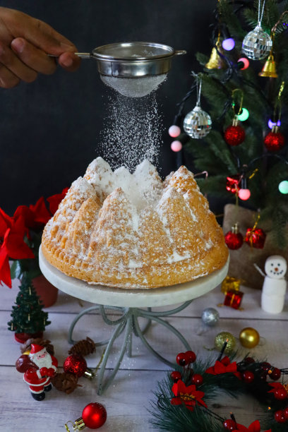 照片中，一个不认识的人在用自制的柠檬细丝制作的Bundt蛋糕上筛糖粉，圣诞树模具，大理石上的柠檬味环形海绵，金属冷却架，筛子，聚焦前景图片下载