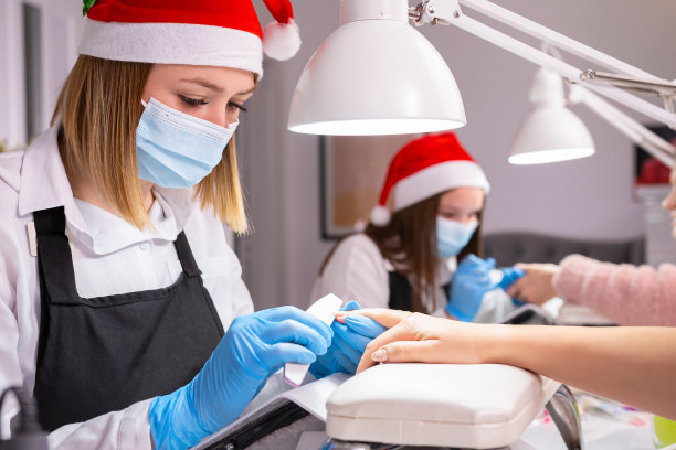 年轻的女专业美甲师在美容院，戴着圣诞帽，戴着医用口罩和防护手套，为一位女士在圣诞节期间做美甲。图片下载