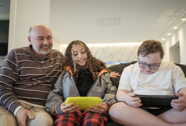 患有唐氏综合症的父亲、女儿和儿子坐在沙发上使用电子平板电脑。图片下载