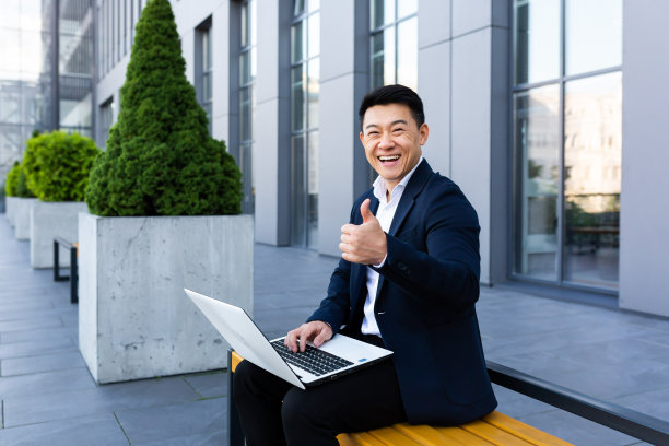 开朗的亚洲男商人正在办公中心附近的电脑上用笔记本电脑进行网上银行业务，面带微笑看着摄像机图片下载