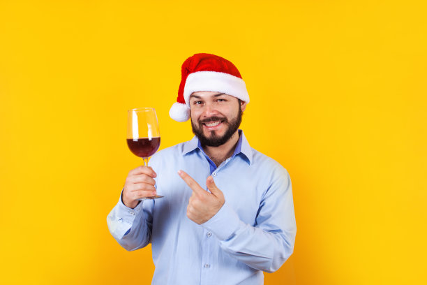 在墨西哥拉丁美洲的黄色背景下，圣诞概念中的拉丁人拿着一杯葡萄酒，戴着圣诞帽，玩得很开心，惊讶地微笑着图片下载