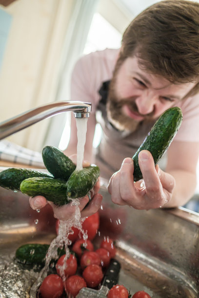 一个一丝不苟的男人在水流下检查并清洗黄瓜、西红柿、辣椒和厨房水槽里的一把刀。图片下载