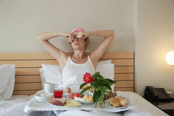 一个戴着粉色睡眠面膜，睡眼惺忪的年轻女人正在床上吃早餐图片下载