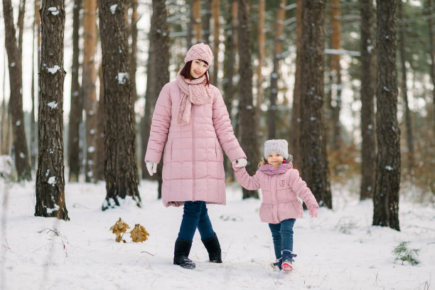 母亲和女儿走在冬天的雪林。快乐的妈妈和穿着粉色外套的小女孩手牵着手，在寒冷的冬日里享受着户外散步的乐趣。图片下载