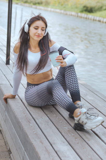 一个穿着运动服的女人在锻炼后戴着耳机听音乐，在河边喝咖啡图片下载