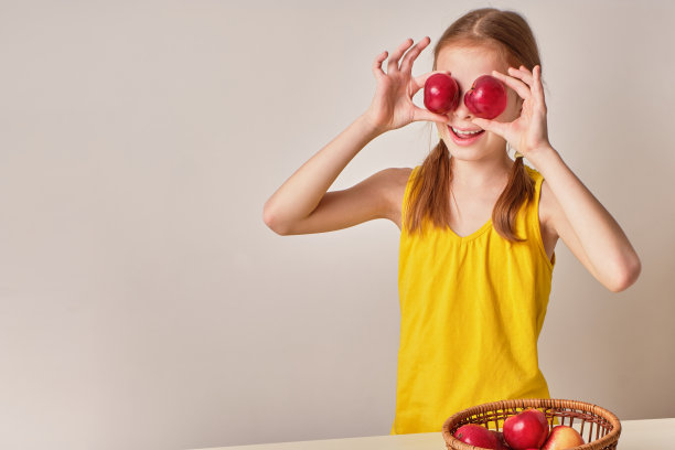 一个积极好玩的女孩与苹果在灰白色的背景。一个快乐的孩子，一个微笑的女孩，手里拿着新鲜的成熟的红苹果。附近有一篮子苹果。健康有机营养的概念图片下载