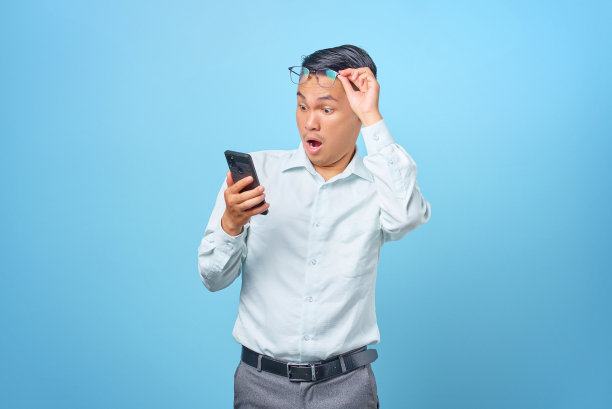 在蓝色背景下，年轻英俊的商人用智能手机摘掉眼镜，震惊了图片下载