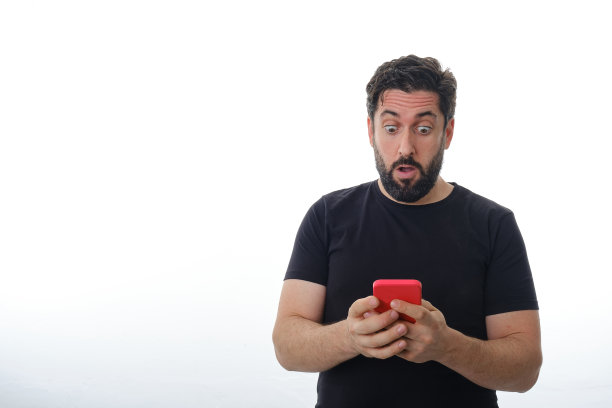 一名男子站在一个孤立的背景上，惊讶地看着自己的手机。图片下载