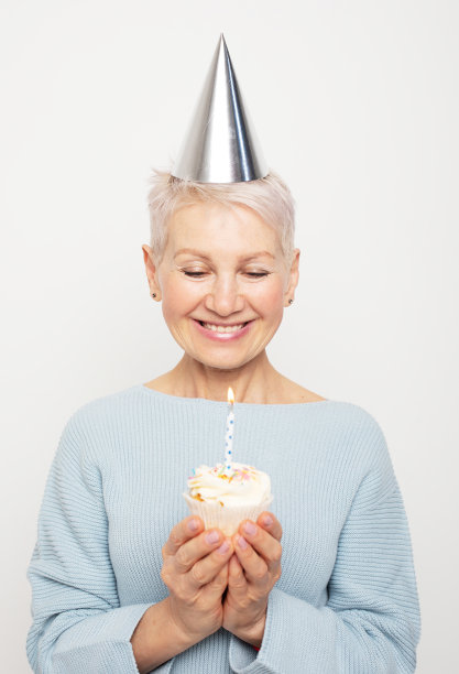 快乐美丽的中年女性戴着圆锥形的帽子，用纸杯蛋糕和蜡烛庆祝生日图片下载