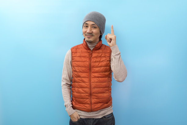 英俊的亚洲男人，戴着帽子，穿着橙色夹克，手指指向白色的孤立背景。图片下载