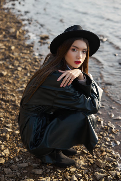 为日落时的年轻女子画像。穿着黑色圆帽和皮革雨衣的时尚女子在湖边。漂亮的妆容和鲜红的唇膏图片下载