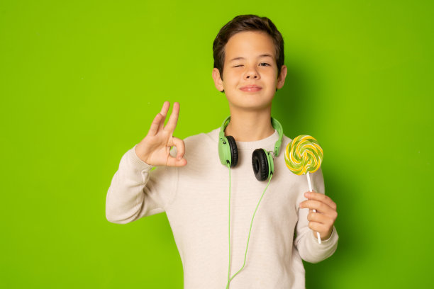 微笑的男孩手里拿着一根棒棒糖，在绿色背景上孤立的ok标志图片下载