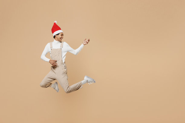 全身冰凉的年轻非洲男子20多岁戴圣诞老人红色圣诞帽跳高演奏吉他手势孤立在素色米色背景工作室肖像新年快乐2022庆祝概念图片下载