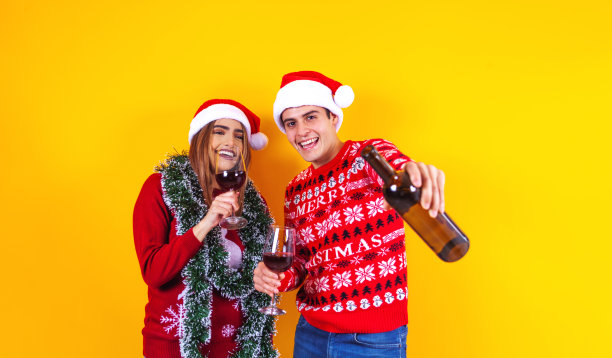 年轻的拉丁夫妇的朋友在墨西哥拉丁美洲，男人和女人穿着红色毛衣圣诞老人帽在黄色背景下拿着酒杯图片下载