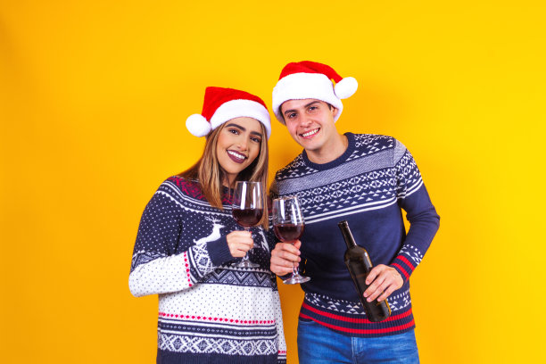年轻的拉丁夫妇的朋友在墨西哥拉丁美洲，男人和女人穿着红色毛衣圣诞老人帽在黄色背景下拿着酒杯图片下载