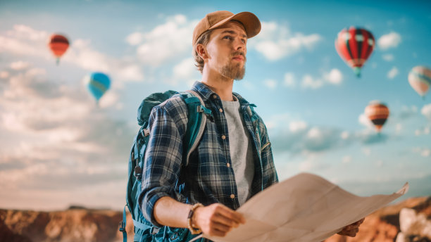 年轻英俊的游客在大自然中背着背包登山。男性冒险背包客在岩石峡谷的顶部用地图导航。国家公园里的热气球。图片下载