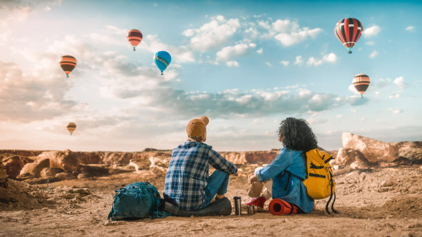 年轻多样的游客夫妇徒步旅行背包，休息在岩石峡谷的顶部。冒险旅行中的男女背包客朋友。国家公园的暮光之城热气球节。图片下载