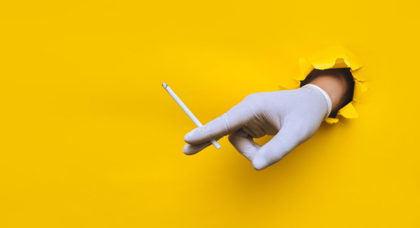 戴着白色保护性医用手套的护士(医生)的手握着一支纤细的香烟。黄色的纸上有个破洞。covid-19期间卫生工作者的吸烟观念和习惯控制图片下载