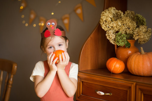 孩子头戴火鸡形状的头饰，背景是感恩节装饰的大南瓜。给孩子们的纸工艺品。为孩子们创造艺术。一个白人女孩的肖像图片下载