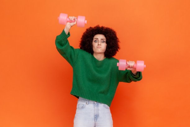 穿着绿色休闲风格毛衣的运动女性，留着非洲式发型，用硬哑铃做手臂运动，表情滑稽。图片下载
