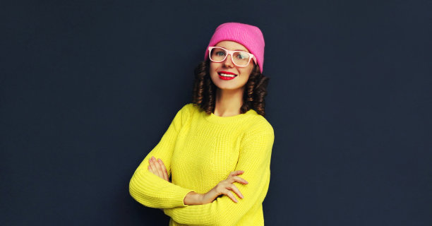 这是一幅时尚微笑的年轻女子的肖像，身穿黄色针织衫，戴着粉红色的帽子，背景为黑色图片下载