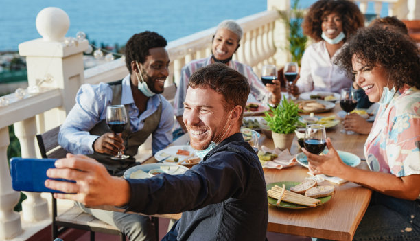 年轻的多种族朋友一起在户外露台上吃晚餐，下巴上戴着外科口罩自拍——冠状病毒和社交距离的概念图片下载