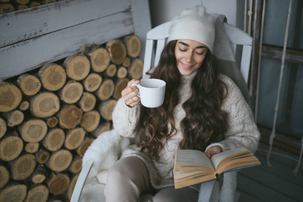 一个年轻漂亮的女人，戴着针织帽，穿着白色毛衣，坐在舒适的房子门廊的扶手椅上喝茶看书。图片下载