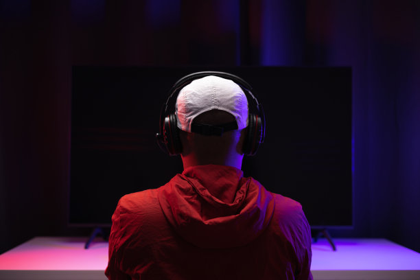 后视图年轻的播放器视频电脑pc。年轻人坐在游戏站的椅子上。Streamer男子戴着耳机在暗室里在线玩动作游戏。图片下载