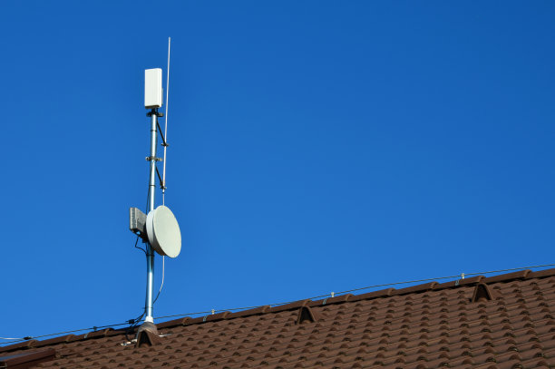 一个互联网信号放大器位于家庭住宅的屋顶上。放大器的抛物线天线和散热片。癌症和脑部疾病的危险。棕色砖瓦山墙屋顶，带通风管道图片下载
