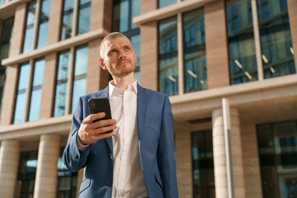 一张低角照片，一位身穿蓝色西装的年轻英俊的金发商人看着别处，想着他刚刚在智能手机上读到的一条信息图片下载