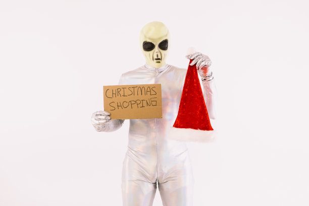 身穿银色套装、戴着绿色外星人面具的人，举着写着“圣诞购物”的牌子，白色背景上戴着圣诞老人的圣诞帽图片下载