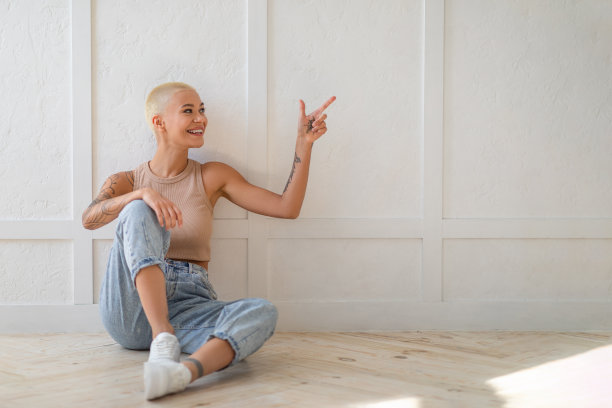 年轻随意的女子坐在白色空墙旁的地板上，指着广告文案空间，自由空间图片下载