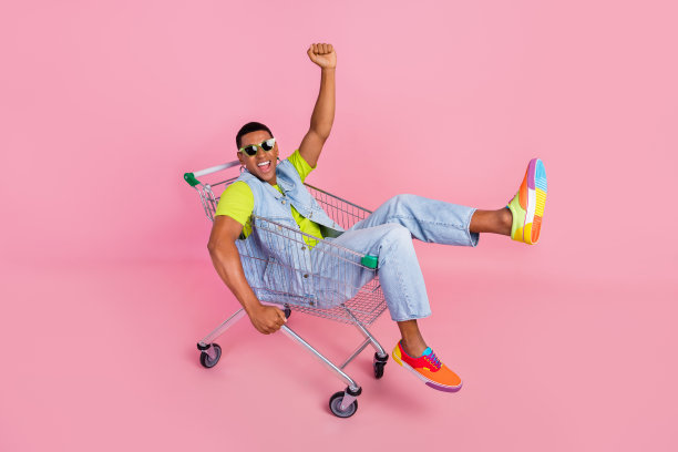 全尺寸照片惊讶的千禧一代深色头发的家伙坐在手推车上，戴着眼镜，t恤，背心，牛仔裤，运动鞋孤立的粉红色背景图片下载
