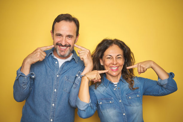 美丽的中年夫妇一起站在孤立的黄色背景下，愉快地微笑，展示和指指点点，牙齿和嘴。牙齿健康的概念。图片下载