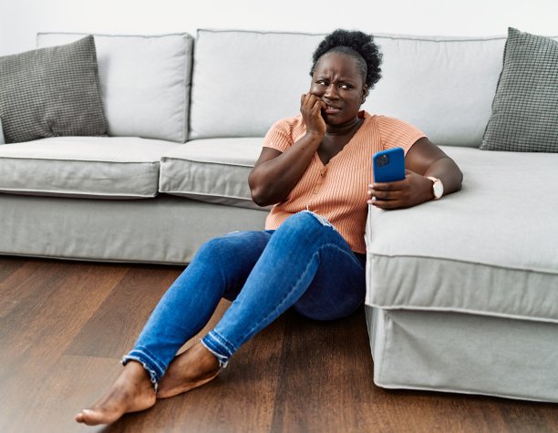 年轻的非洲妇女坐在家里的地板上使用智能手机，看起来紧张和紧张，手放在嘴里咬着指甲。焦虑的问题。图片下载