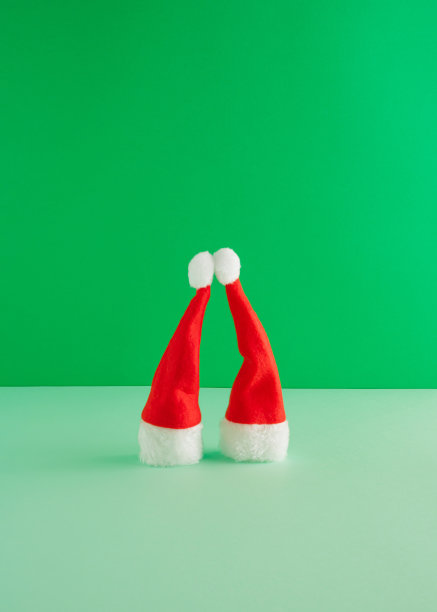 红色圣诞帽在鲜艳的绿色背景上的创造性安排。最小的新年概念。一对情侣接吻的灵感。图片下载