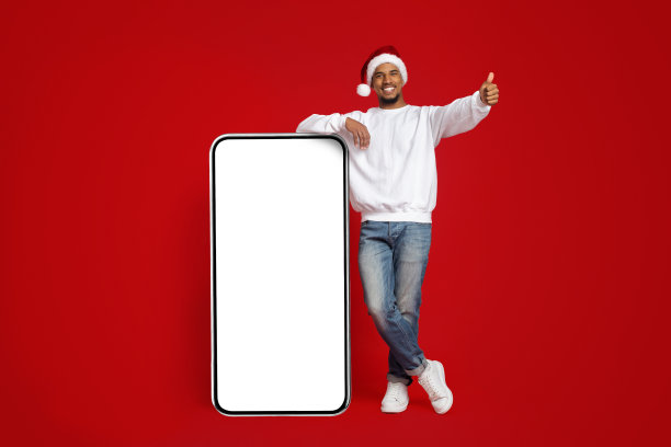 戴着圣诞帽的快乐男人站在巨大的智能手机模型旁图片下载