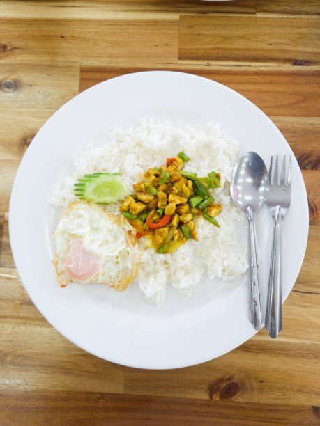 咖喱鸡炒酱，扁豆拌饭，黄瓜和煎蛋。一个白色的盘子放在一张木桌上。有不锈钢的勺子和叉子。图片下载