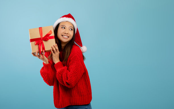 快乐的亚洲女士戴着圣诞帽，拿着礼物，摇晃着破碎的盒子，看着蓝色背景上的自由空间图片下载