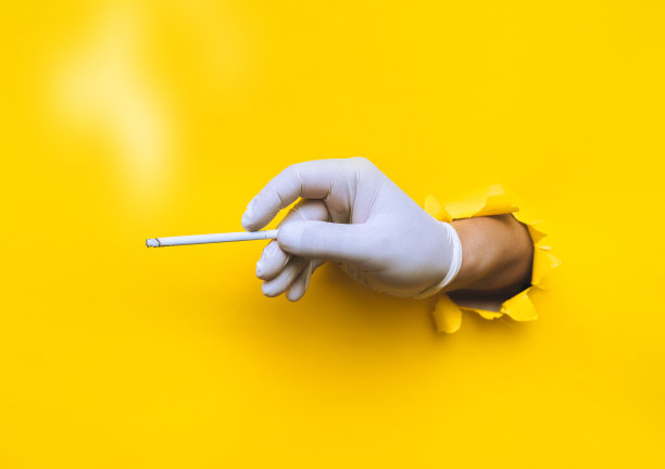 戴着白色乳胶医用手套的医生(护士)的手握着一支纤细的香烟。黄色的纸上有个破洞。covid-19期间卫生工作者的吸烟观念和习惯控制图片下载