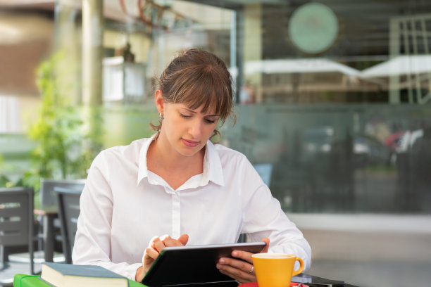 一位女商人在咖啡馆里用她的电子平板电脑，桌上放着一杯咖啡、一些书和文件夹图片下载