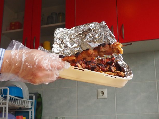 一次性包装的烤肉和餐厅的铝箔。从餐馆和咖啡馆送货到家。女性的手戴着一次性手套，用棍子拿着肉。红色家具厨房图片下载
