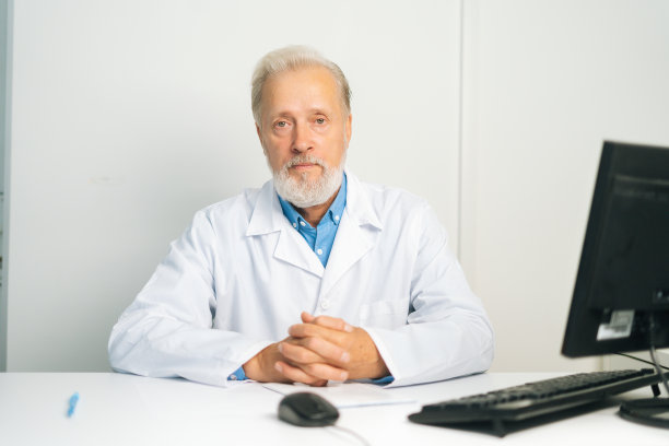 照片中，严肃成熟的成年男医生穿着白色的医疗服，坐在电脑旁，看着摄像机。图片下载