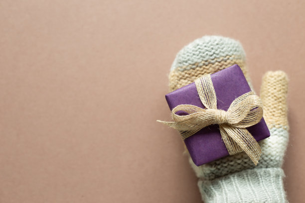 手在冬季针织手套与紫色礼品盒上的棕色背景。周年纪念，圣诞节，问候概念。俯视图，复制空间图片下载