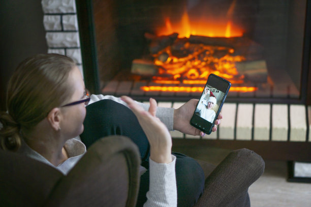 照片中，一位中年妇女坐在壁炉旁，手里拿着手机，微笑着打视频电话或自拍。通过视频链接祝贺圣诞快乐的概念。图片下载