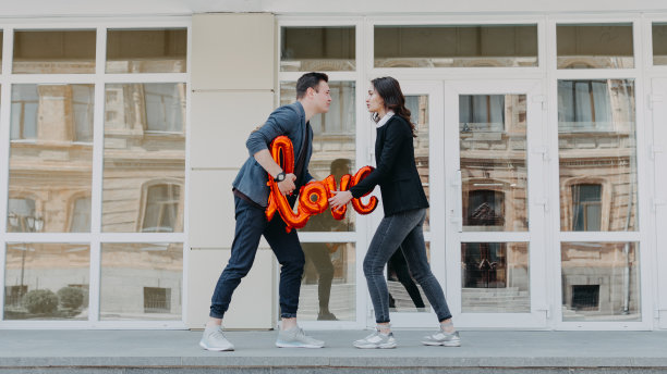 情人节庆祝和约会的概念。幸福的情侣在城市的街道上拿着红色的爱情气球。户外照片的年轻夫妇与形状的气球字爱。图片下载