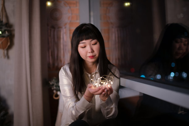 一位年轻美丽的亚裔韩国女性在自己舒适的房子里拿着点亮的节日花环，在窗户上做着晚上积极情绪的梦。新年和圣诞节的概念。图片下载